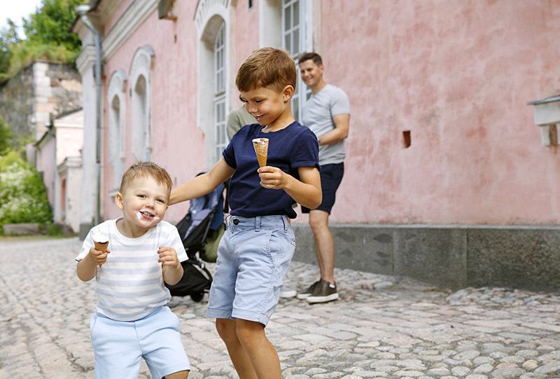 Zwei Jungs mit Eiscreme in der Hand albern miteinander herum. Im Hintergrund steht der Vater mit Kinderwagen.