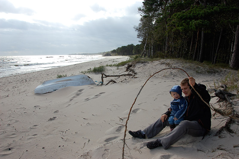 Ein Vater sitzt mit seinem Baby am Strand und hat einen großen Stock in der Hand.