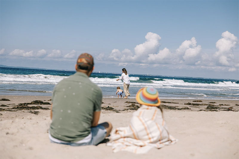 Eine Familie sitzt am Strand. Vater und Kind sitzen nebeneinander und beobachten, wie Mutter und Kinder ins Wasser laufen.