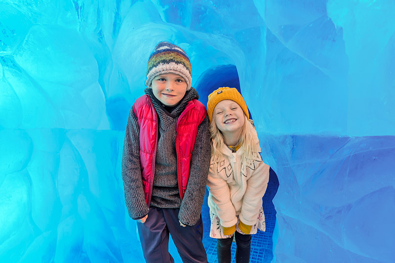 Zwei Kinder stehen vor einer eisblauen Wand in Winterkleidung