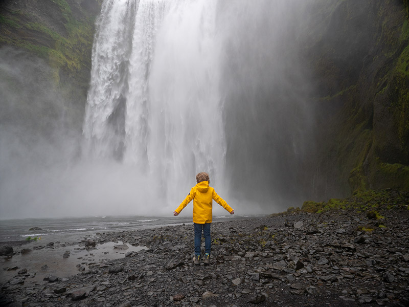 Ein Junge mit gelber Regenjacke steht am Fuße eines Wasserfalls mit ausgebreiteten Armen.