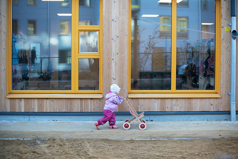 Ein Kleinkind schiebt einen Spielzeug-Kinderwagen vor sich her vor einem großen Schaufenster.