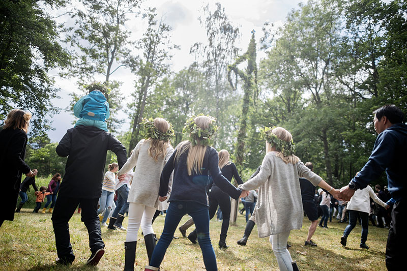 Erwachsene und Kinder tanzen Hand in Hand um eine Maistange und tragen Blumenkränze im Haar.