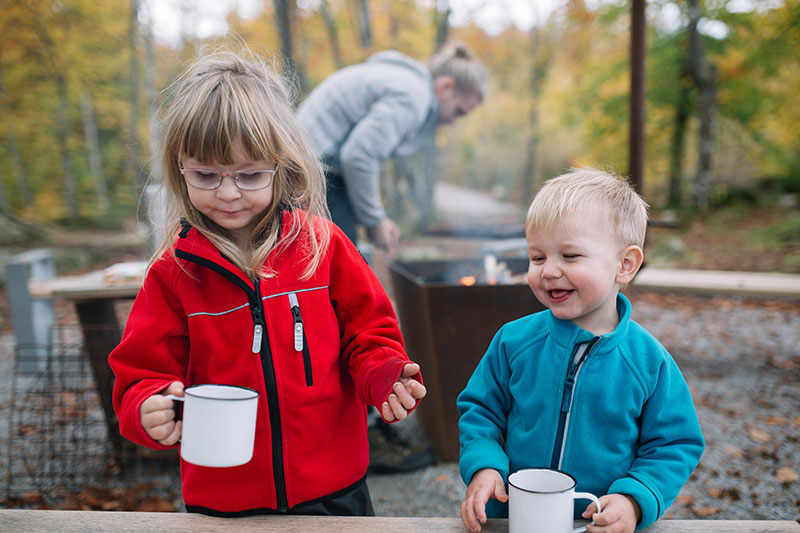 Zwei Kinder stehen mit Campingtassen im Wald. Im Hintergrund steht ein Mann am Feuer.