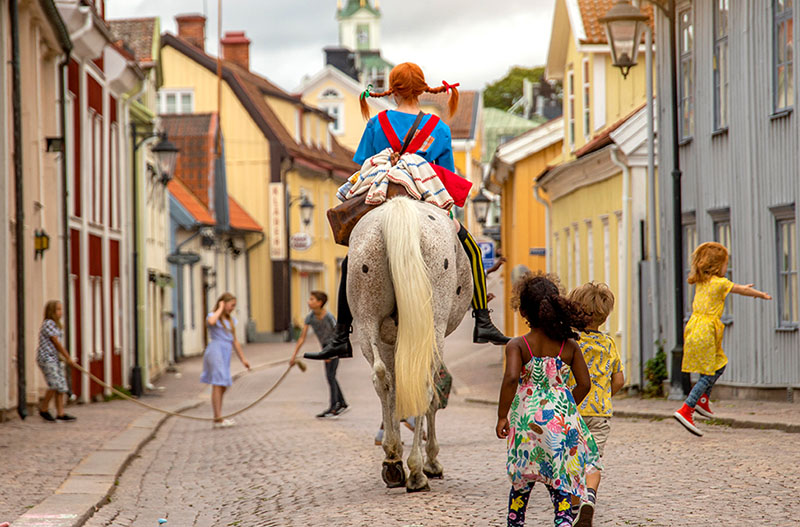 Pippi reitet auf einem weißen Pferd über eine Straße in der Astrid Lindgrens Welt. Um sie herum spielen mehrere Kinder.