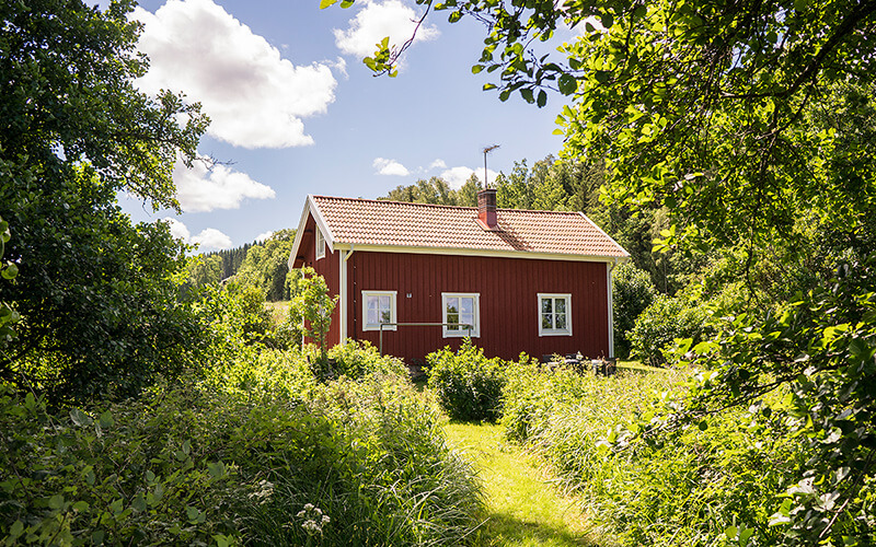 Rotes Holzhaus mit Satteldach umgeben von dichtem Grün und Bäumen unter blauem Himmel mit Wolken.