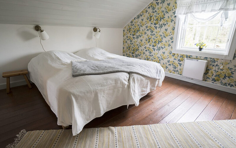 Schlafzimmer mit zwei Einzelbetten unter einer Dachschräge, weißen Bettdecken, einer Bank an der Wand und einer Fensterfront mit floral gemusterter Tapete.