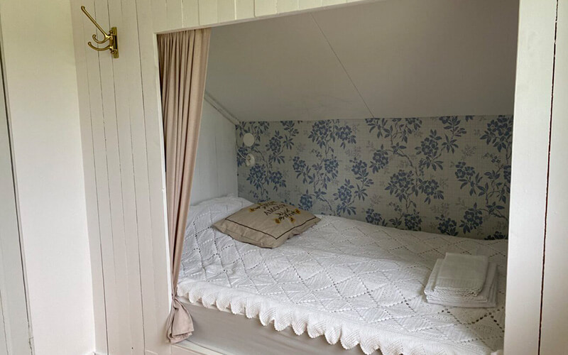 Gemütliche Nische mit schräger Decke, weißer Wandverkleidung, floral gemusterter Tapete, weißer gehäkelter Tagesdecke, Kissen und gefalteten Handtüchern.