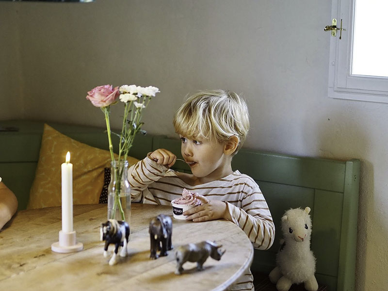 Ein blonder Junge mit gestreiftem Pulli sitzt auf einer grünen Holzbank und isst ein Eis.
