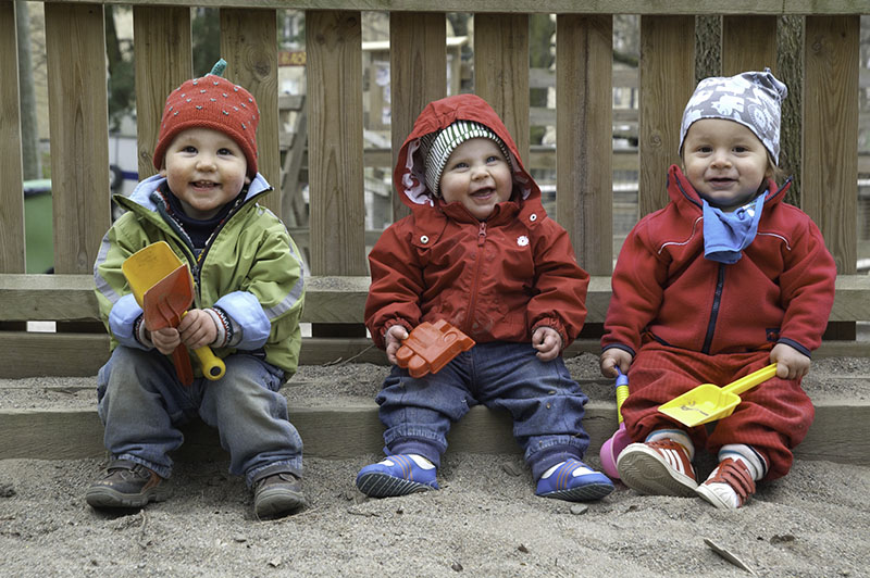 Drei kleine Babys in bunter Kleidung sitzen im Sandkasten und strahlen in die Kamera.
