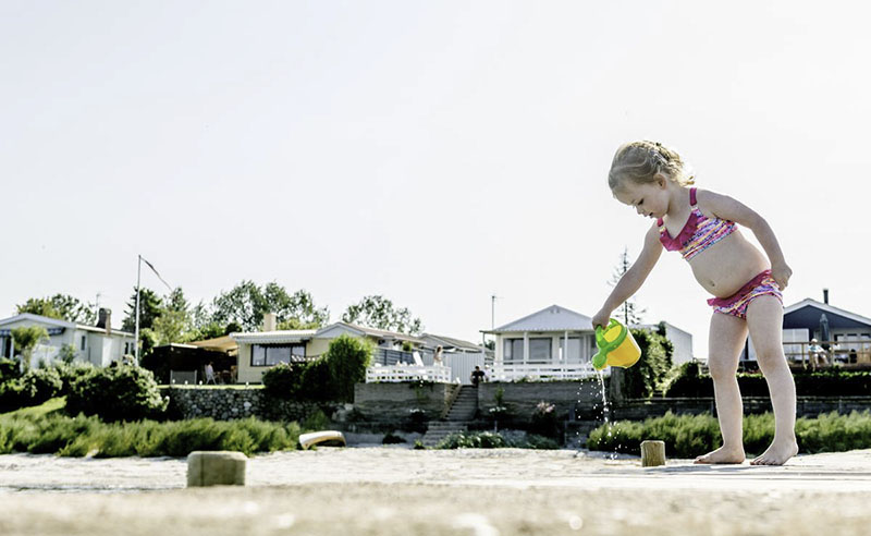 Ein kleines Mädchen in einem pinken Bikini spielt mit einer gelben Gießkanne am Strand.