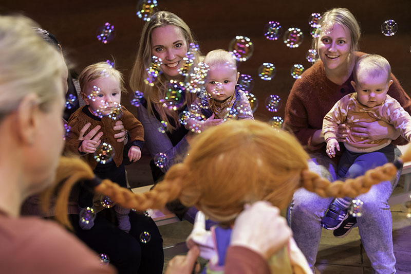 Im Vordergrund ist eine Pippi Langstrumpf Handpuppe. Im Hintergrund sieht man Babys auf den Schößen ihrer Mütter, die die Puppe neugierig ansehen. Überall fliegen Seifenblasen herum.