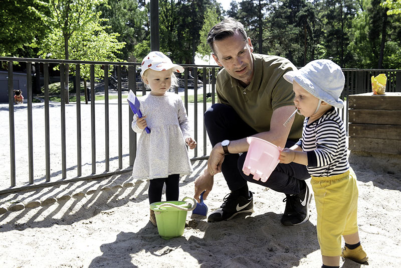 Ein Mann kniet neben zwei Babys im Sandkasten. Beide Babys haben Sandspielzeug in den Händen.